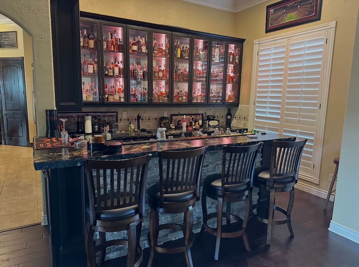 Home bar with backlit liquor display, dark cabinetry, stone front and granite top